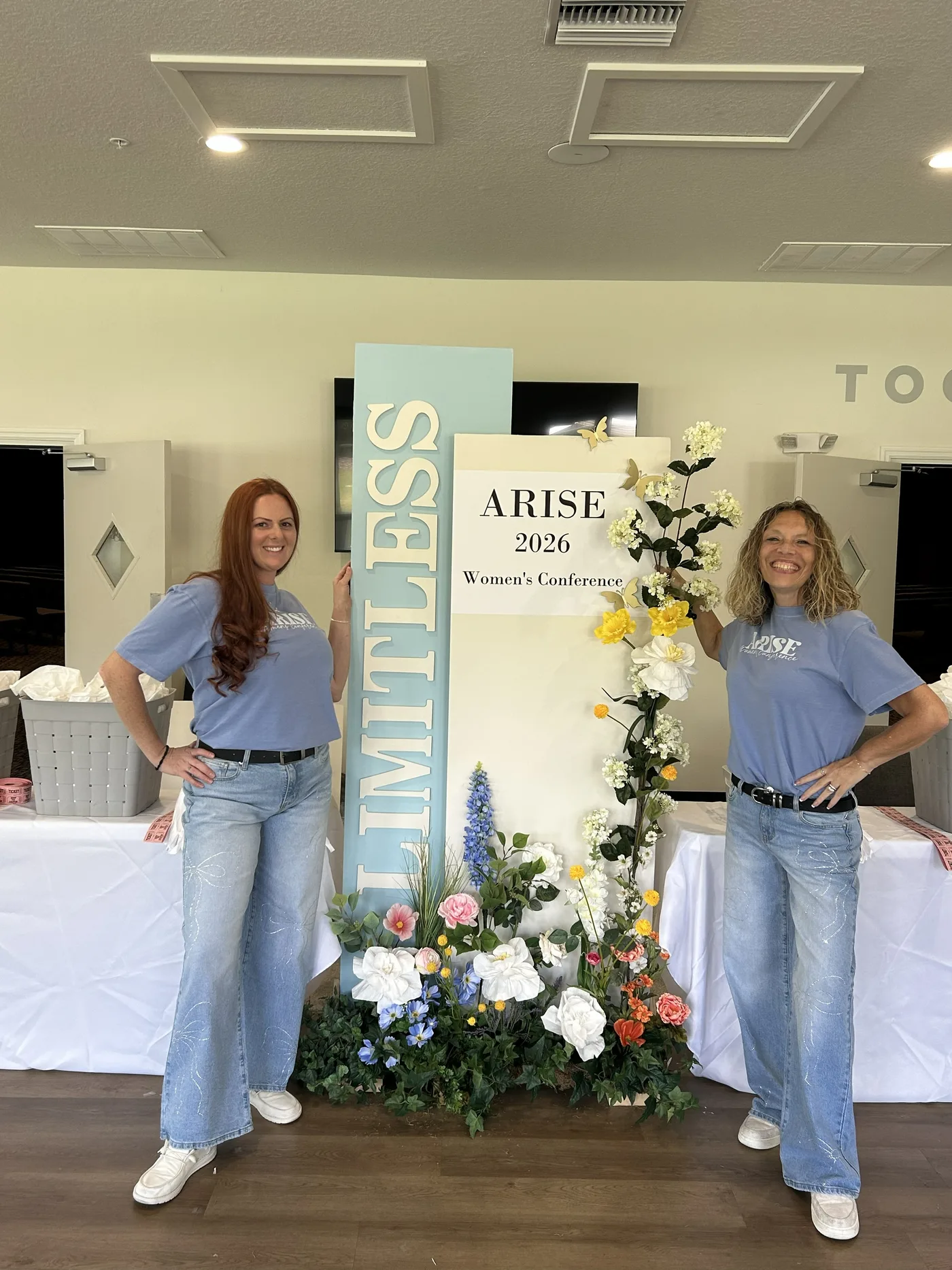 Angela and her daughter Kimmy standing either side of a floral ARISE 2026 Women’s Conference display, both smiling.